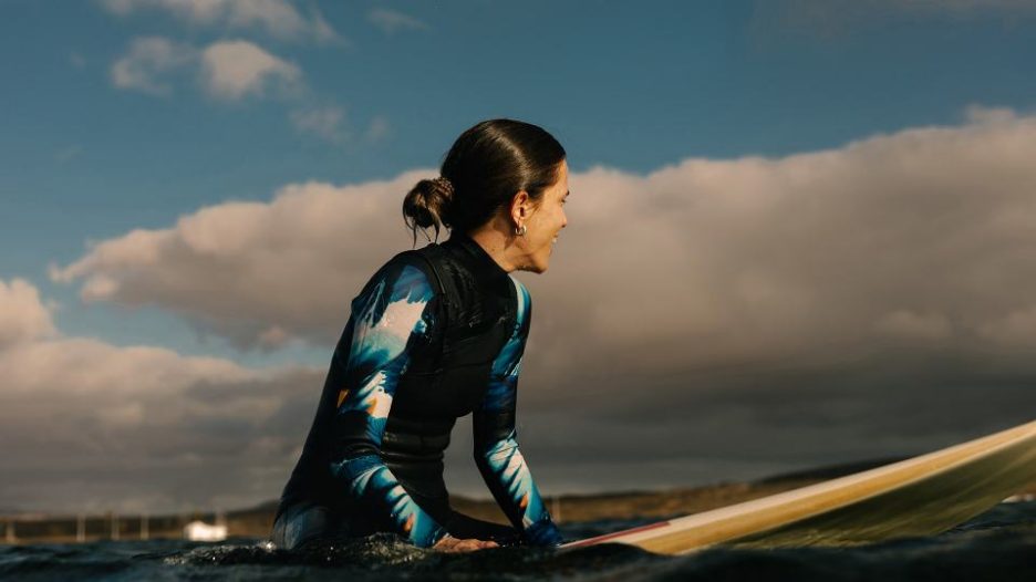 a female surfer patiently awaits the perfect swell, floating on her surfboard while admiring the sunset over the horizon