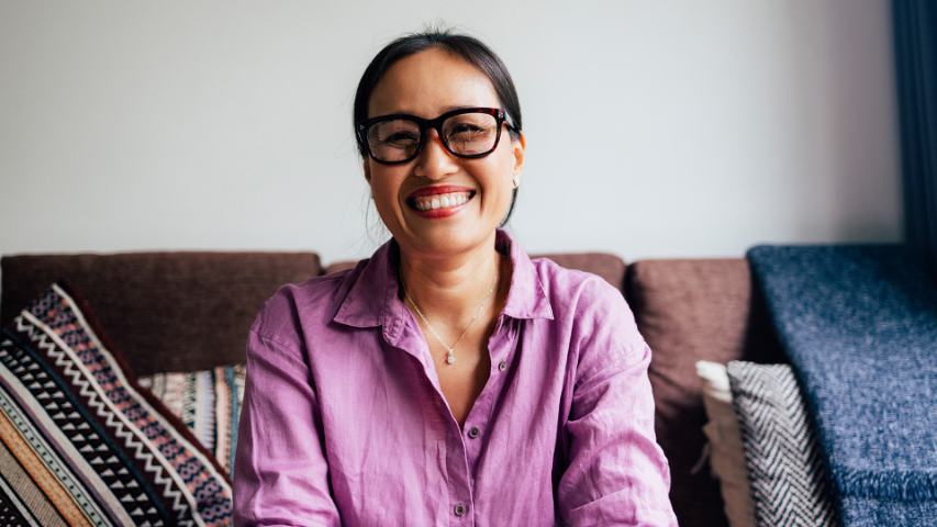 a happy woman in a pink shirt faces the camera as she sits on a brown sofa