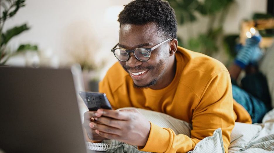 man with glasses is smilng while using his phone and laptop in his room
